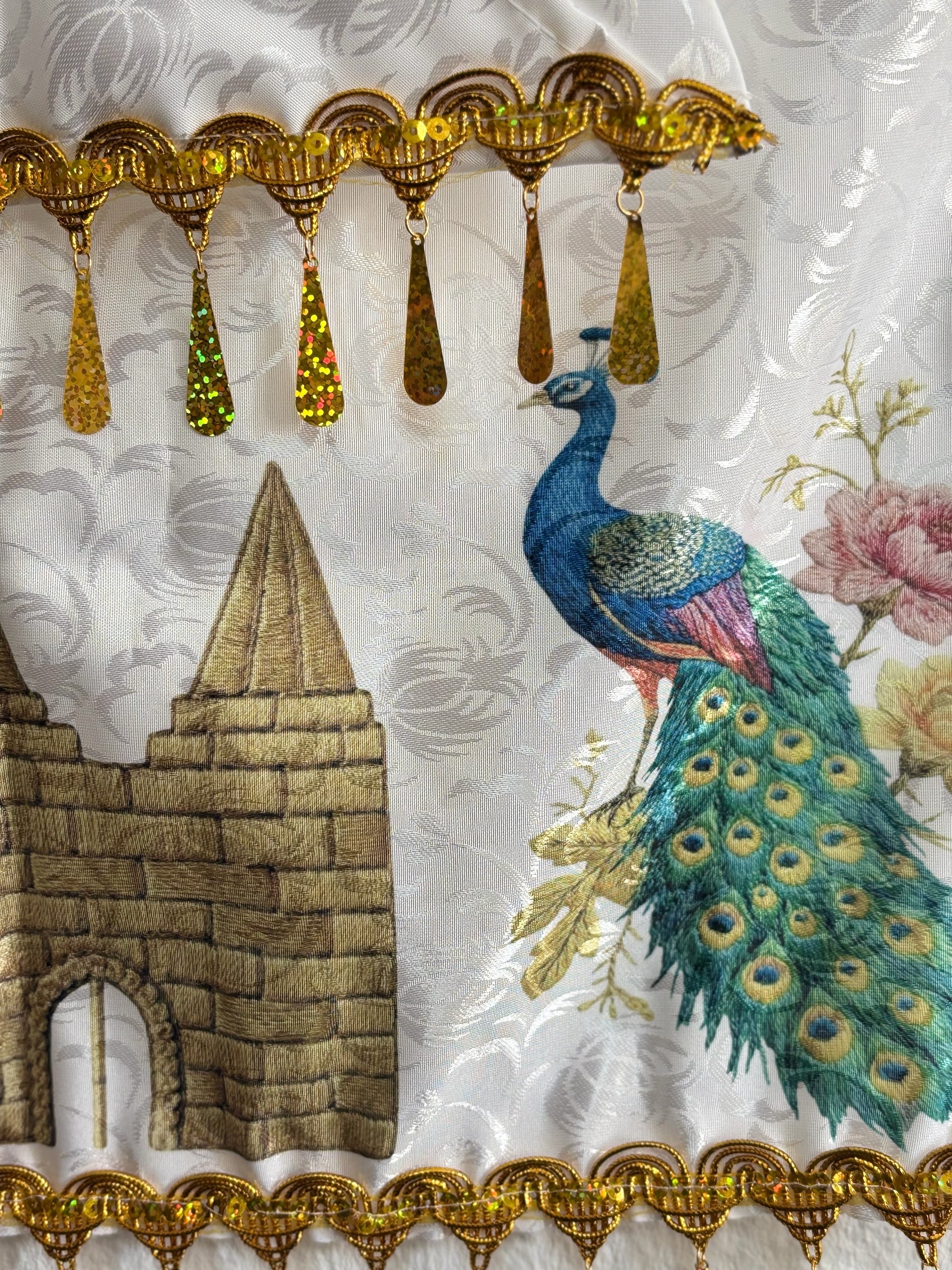 Milan Geschenk e.K Wandbehang mit Pfauenmotiv, goldenen Pailletten und tropfenförmigen Anhängern auf weißem Textil.